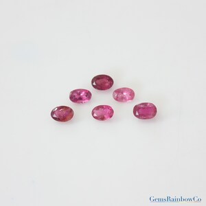 May include: Six oval-shaped pink gemstones of varying shades, arranged in a triangular pattern on a white surface.