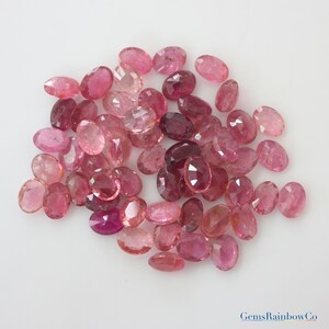May include: A collection of pink oval-shaped gemstones. The gemstones are various shades of pink, from light pink to deep pink. The gemstones are arranged in a pile on a white background.