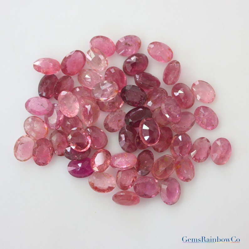 May include: A collection of pink oval-shaped gemstones. The gemstones are various shades of pink, from light pink to deep pink. The gemstones are arranged in a pile on a white background.