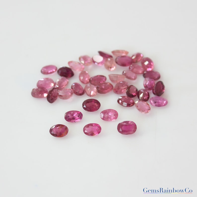 May include: A collection of pink and red oval-shaped gemstones. The gemstones are scattered on a white surface.