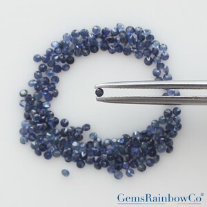May include: A collection of small, round, faceted dark blue sapphires arranged in a circular pattern, with a single matching sapphire held by a pair of silver-toned metal tweezers. The text GemsRainbowCo is visible in the bottom right corner.
