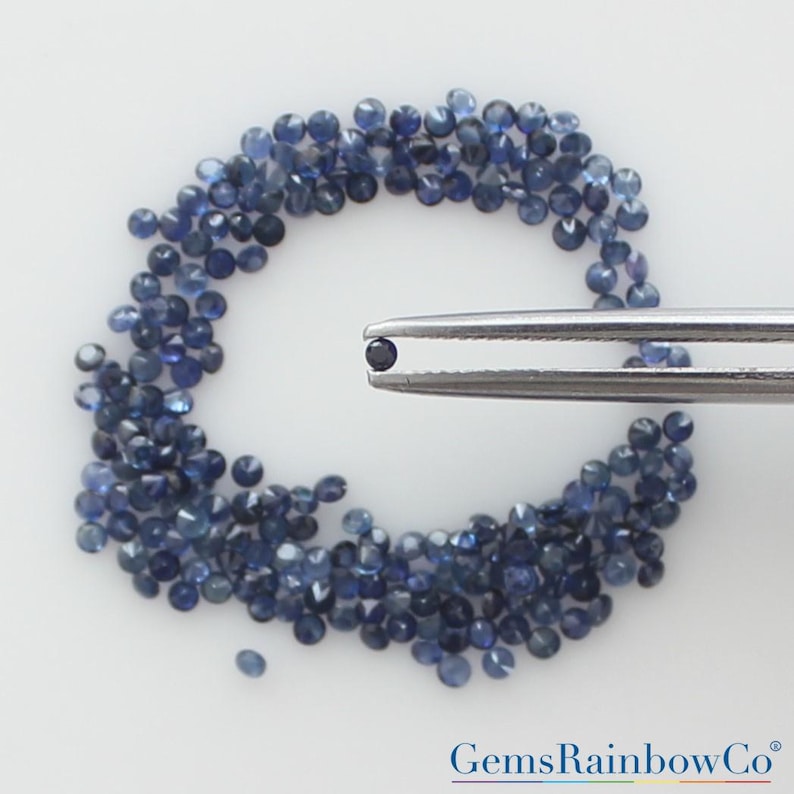 May include: A collection of small, round, faceted dark blue sapphires arranged in a circular pattern, with a single matching sapphire held by a pair of silver-toned metal tweezers. The text GemsRainbowCo is visible in the bottom right corner.