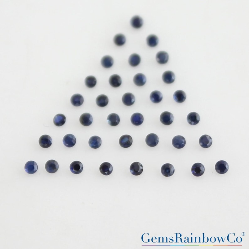 May include: A collection of small, round, faceted dark blue gemstones arranged in a triangular formation. The stones are uniform in shape and color, displaying a deep navy hue with reflective surfaces. The text GemsRainbowCo is visible.