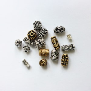 Könnte beinhalten: Eine Sammlung von silbernen und goldenen Metallperlen mit komplizierten Designs. Die Perlen sind in verschiedenen Formen und Größen, darunter rund, oval und zylindrisch.