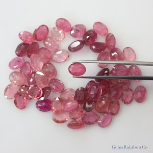 May include: A collection of pink oval-shaped gemstones, some with a slight reddish hue, arranged in a circle on a white surface. One gemstone is held by tweezers in the center of the circle.