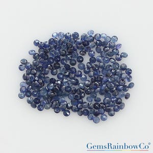May include: A collection of numerous small, round, faceted blue sapphires arranged in a loose cluster. Each gemstone displays a deep navy to light blue hue with reflective surfaces. The text GemsRainbowCo is printed in the bottom right corner.