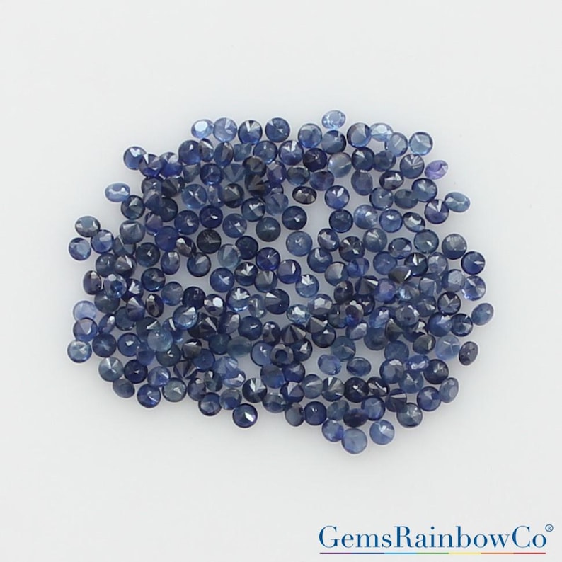 May include: A collection of numerous small, round, faceted blue sapphires arranged in a loose cluster. Each gemstone displays a deep navy to light blue hue with reflective surfaces. The text GemsRainbowCo is printed in the bottom right corner.
