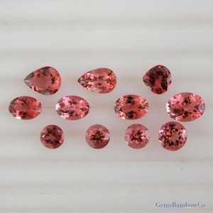 May include: Ten loose pink tourmaline gemstones of various shapes and sizes. The gemstones are arranged on a white surface.