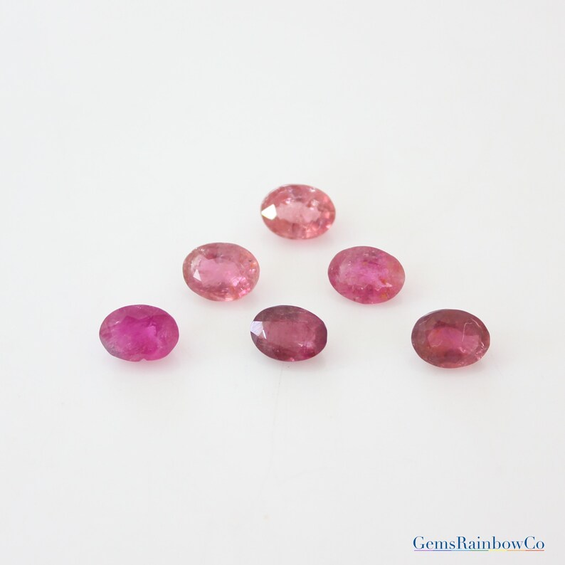 May include: Six oval-shaped pink gemstones, likely tourmalines, on a white background. The gemstones are various shades of pink, from light to dark.