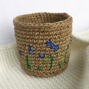 May include: A cylindrical basket crafted from natural brown jute twine, featuring a hand-embroidered pattern of blue flowers with green stems and leaves encircling the exterior. The basket has a sturdy, woven texture and an open top design.