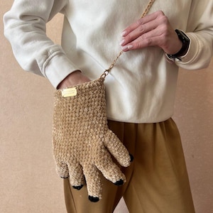 Peut inclure: Un sac à main original en forme de main, de couleur beige, avec une chaîne dorée. Le sac est fait d'un matériau doux et texturé, avec des détails noirs sur le bout des doigts. Une petite étiquette dorée est attachée.