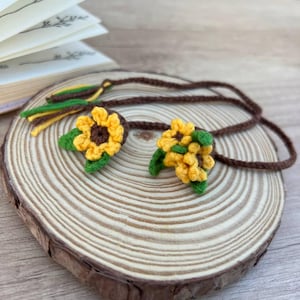 Op de afbeelding: Een bruin gehaakt koord met twee zonnebloemmotieven met gele bloemblaadjes, donkerbruine harten en groene bladeren. Het koord eindigt in een kwastje met gele en groene garenstrengen. Het item ligt op een ronde houten schijf.