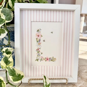 May include: A framed floral art print featuring the letter "L" adorned with watercolor flowers and two small bees. The white frame has a pink and white striped border. The print is displayed on a decorative gold stand.