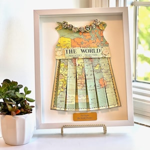 May include: A framed world map in the shape of a dress with a gold trim and a yellow tag that reads "For Taylor, The World Awaits..."