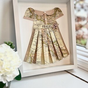Puede incluir: Un marco blanco con un mapa de Italia hecho a la forma de un vestido. El vestido tiene un cuello rosa y un cierre de bot&oacute;n. El vestido est&aacute; decorado con una flor rosa.