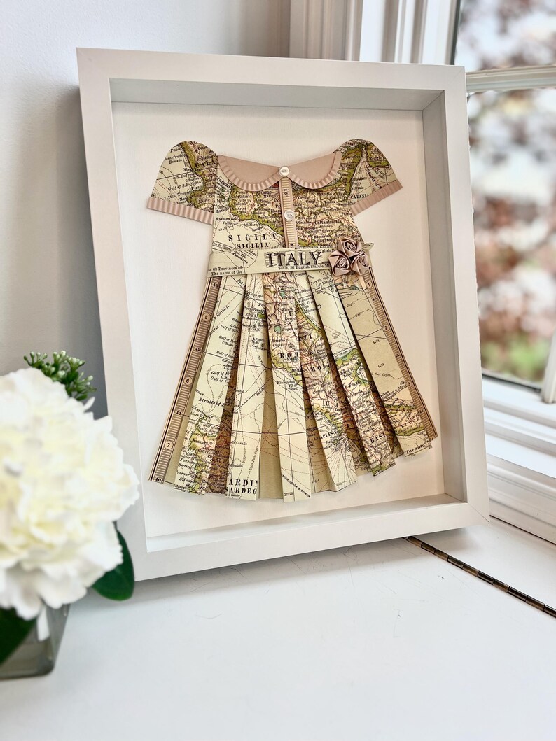 Puede incluir: Un marco blanco con un mapa de Italia hecho a la forma de un vestido. El vestido tiene un cuello rosa y un cierre de bot&oacute;n. El vestido est&aacute; decorado con una flor rosa.