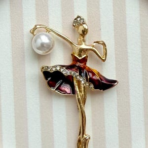 May include: A gold-toned ballerina brooch featuring a dark red enamel skirt accented with clear rhinestones. The figure holds a single white faux pearl in one raised arm and wears a small rhinestone headpiece on a smooth gold-toned head.