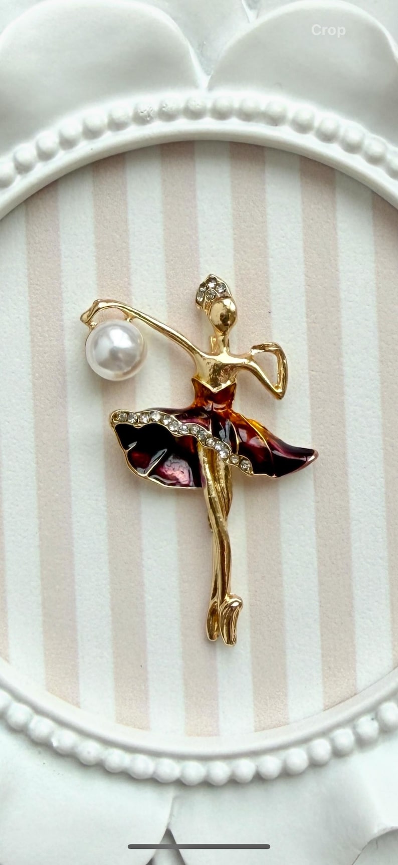 May include: A gold-toned ballerina brooch featuring a dark red enamel skirt accented with clear rhinestones. The figure holds a single white faux pearl in one raised arm and wears a small rhinestone headpiece on a smooth gold-toned head.