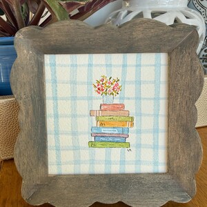 May include: A framed watercolor painting featuring a stack of colorful books topped with a vase of flowers. The background is a blue and white checkered pattern. The frame is a scalloped edge, weathered gray wood.