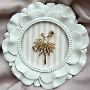 May include: A gold-toned brooch shaped like a ballerina with a white bodice and a skirt embellished with clear crystals. The figure is displayed inside a circular white frame with a scalloped edge and a light pink and white striped background.