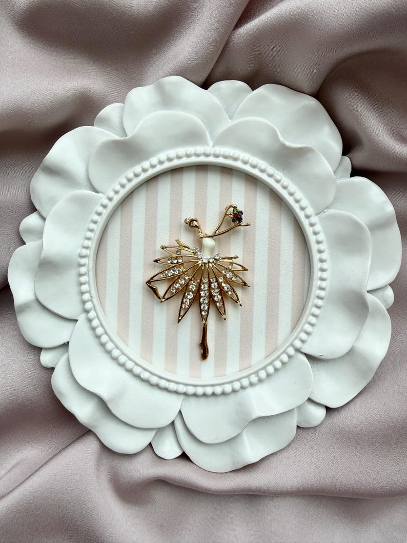 May include: A gold-toned brooch shaped like a ballerina with a white bodice and a skirt embellished with clear crystals. The figure is displayed inside a circular white frame with a scalloped edge and a light pink and white striped background.