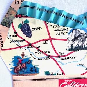 Pu&ograve; includere: Grembiule fatto a mano con un design vintage della mappa della California. Il grembiule ha un bordo a balze azzurro e spalline rosa. La mappa include illustrazioni di uva, del Parco Nazionale di Yosemite e un cowboy a cavallo.