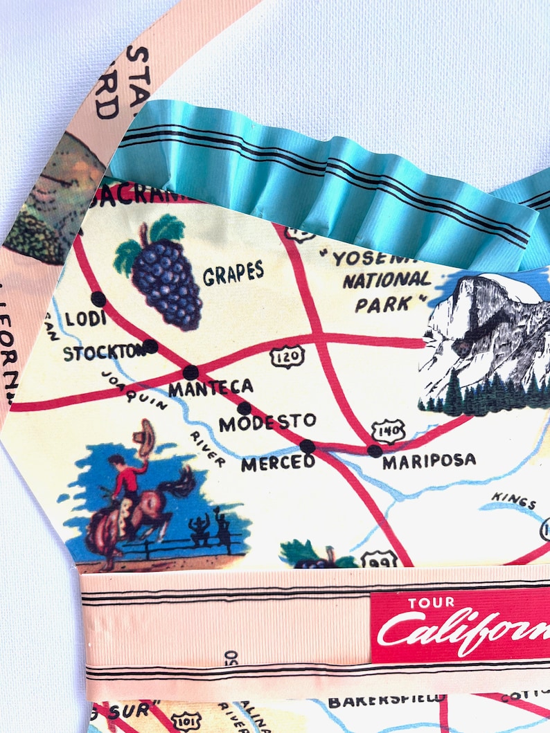 Pu&ograve; includere: Grembiule fatto a mano con un design vintage della mappa della California. Il grembiule ha un bordo a balze azzurro e spalline rosa. La mappa include illustrazioni di uva, del Parco Nazionale di Yosemite e un cowboy a cavallo.