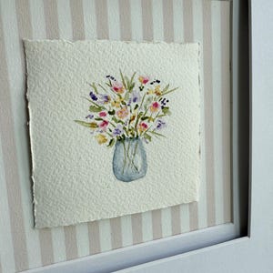 Pu&ograve; includere: Dipinto ad acquerello di un bouquet di fiori in un vaso blu. L'opera d'arte &egrave; su carta ruvida e incorniciata in una cornice bianca. Lo sfondo presenta un motivo a righe nei toni del rosa e del bianco.