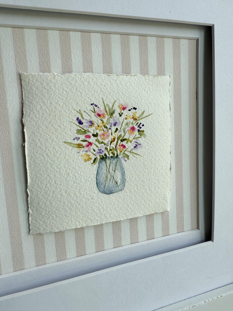 Pu&ograve; includere: Dipinto ad acquerello di un bouquet di fiori in un vaso blu. L'opera d'arte &egrave; su carta ruvida e incorniciata in una cornice bianca. Lo sfondo presenta un motivo a righe nei toni del rosa e del bianco.