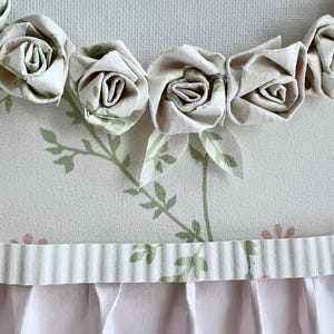 May include: A white fabric background with a row of white fabric roses along the top edge. The background has a green floral pattern and a white ruffled trim along the bottom edge.