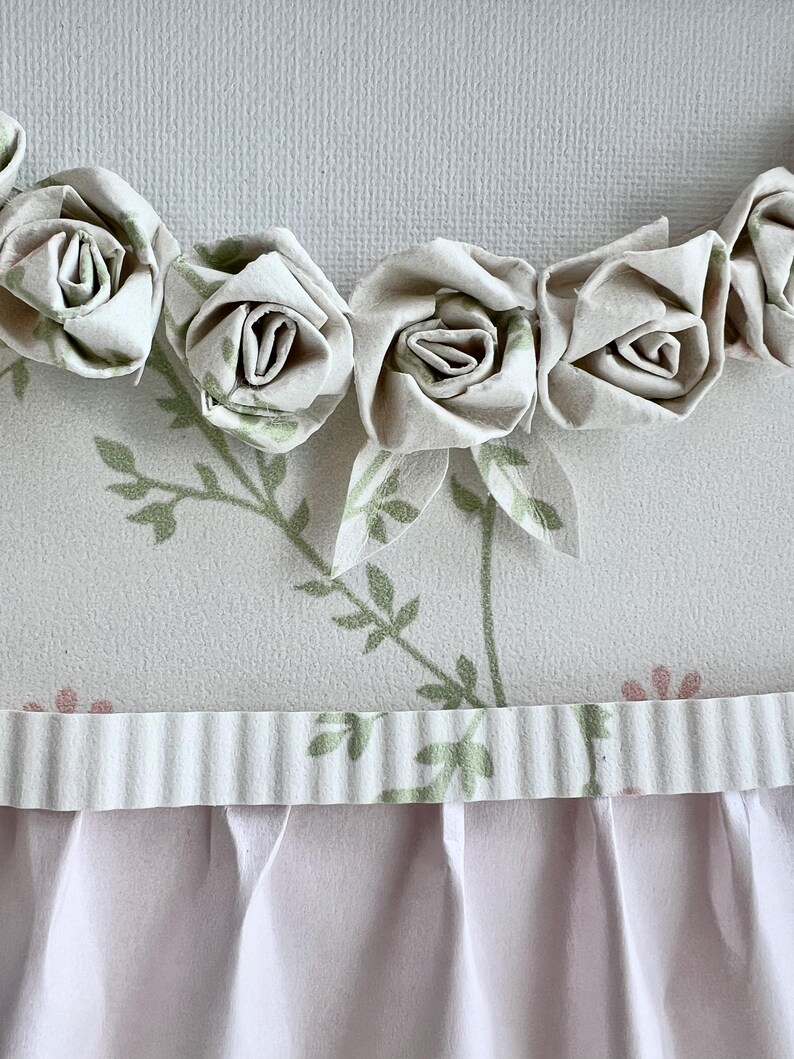 May include: A white fabric background with a row of white fabric roses along the top edge. The background has a green floral pattern and a white ruffled trim along the bottom edge.