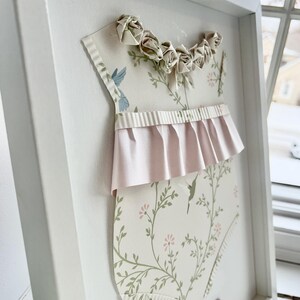 May include: A white frame with a pink and green baby onesie cutout. The onesie has a floral pattern and a pink ruffle trim. The frame has a gold nameplate with the text "Taylor James June 10, 2023".