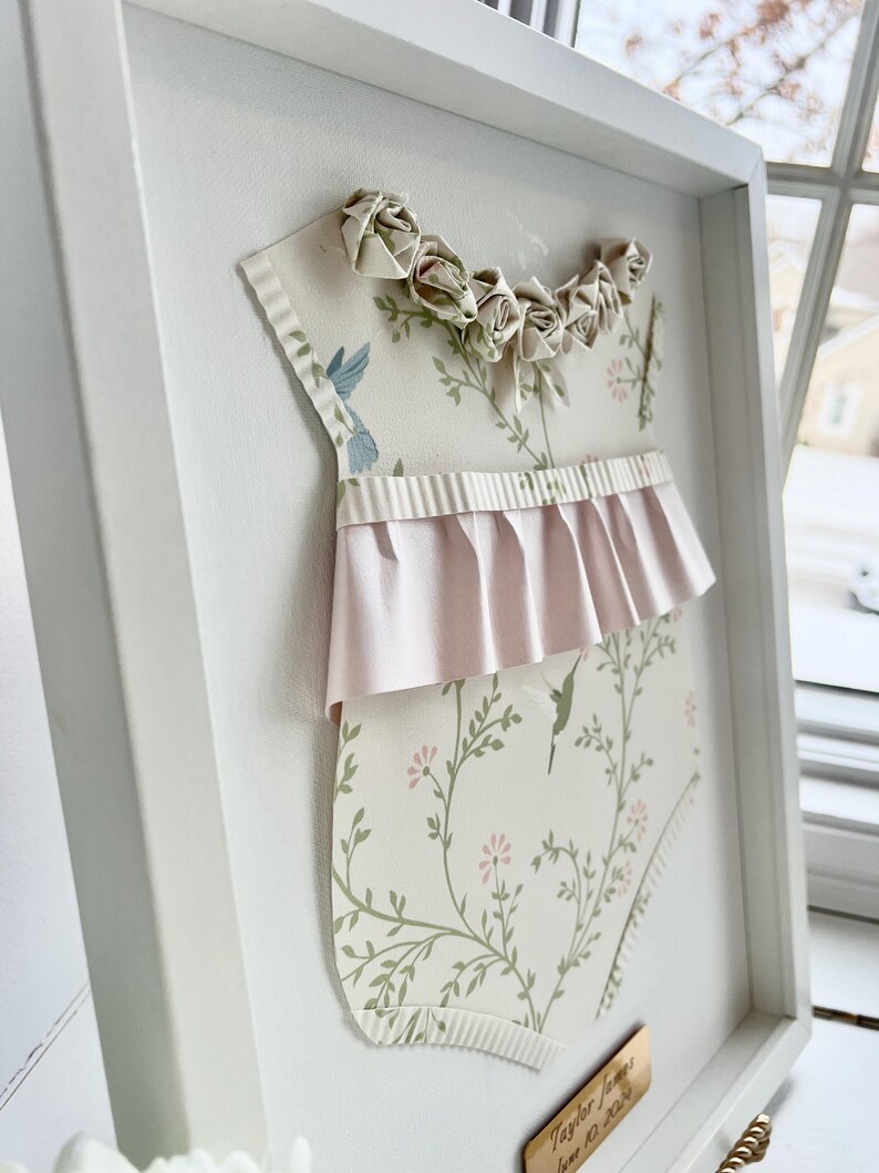 May include: A white frame with a pink and green baby onesie cutout. The onesie has a floral pattern and a pink ruffle trim. The frame has a gold nameplate with the text "Taylor James June 10, 2023".