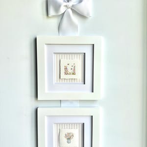 Pu&ograve; includere: Due stampe artistiche floreali incorniciate con cornici bianche e un nastro bianco. L'arte presenta dipinti ad acquerello di fiori e api. Le cornici sono quadrate e montate su un muro bianco.