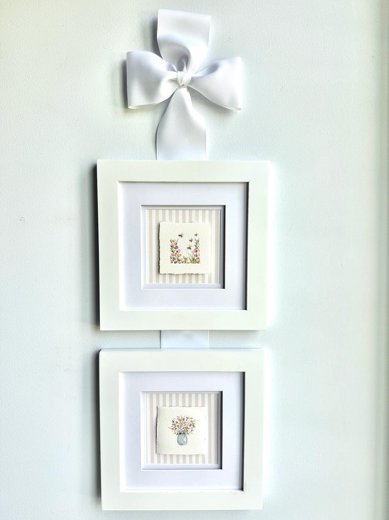 Pu&ograve; includere: Due stampe artistiche floreali incorniciate con cornici bianche e un nastro bianco. L'arte presenta dipinti ad acquerello di fiori e api. Le cornici sono quadrate e montate su un muro bianco.