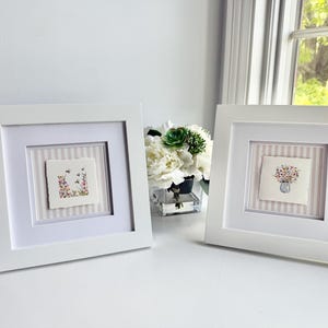 Pu&ograve; includere: Due opere d'arte quadrate con cornice bianca. Ognuna presenta un acquerello su carta ruvida. Uno mostra fiori e api, l'altro un bouquet in un vaso. Entrambi sono su uno sfondo a righe.