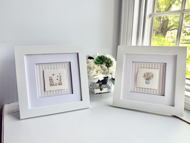 Pu&ograve; includere: Due opere d'arte quadrate con cornice bianca. Ognuna presenta un acquerello su carta ruvida. Uno mostra fiori e api, l'altro un bouquet in un vaso. Entrambi sono su uno sfondo a righe.