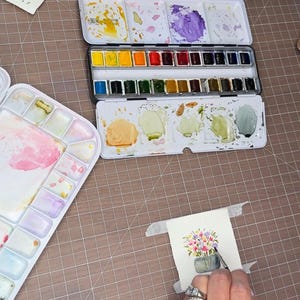 Pu&ograve; includere: Un'attrezzatura per la pittura ad acquerello con una tavolozza di metallo con vari colori ad acquerello e una tavolozza di plastica separata. Si sta lavorando a un piccolo dipinto ad acquerello di fiori in un vaso con un pennello. L'attrezzatura &egrave; su una superficie a griglia marrone.