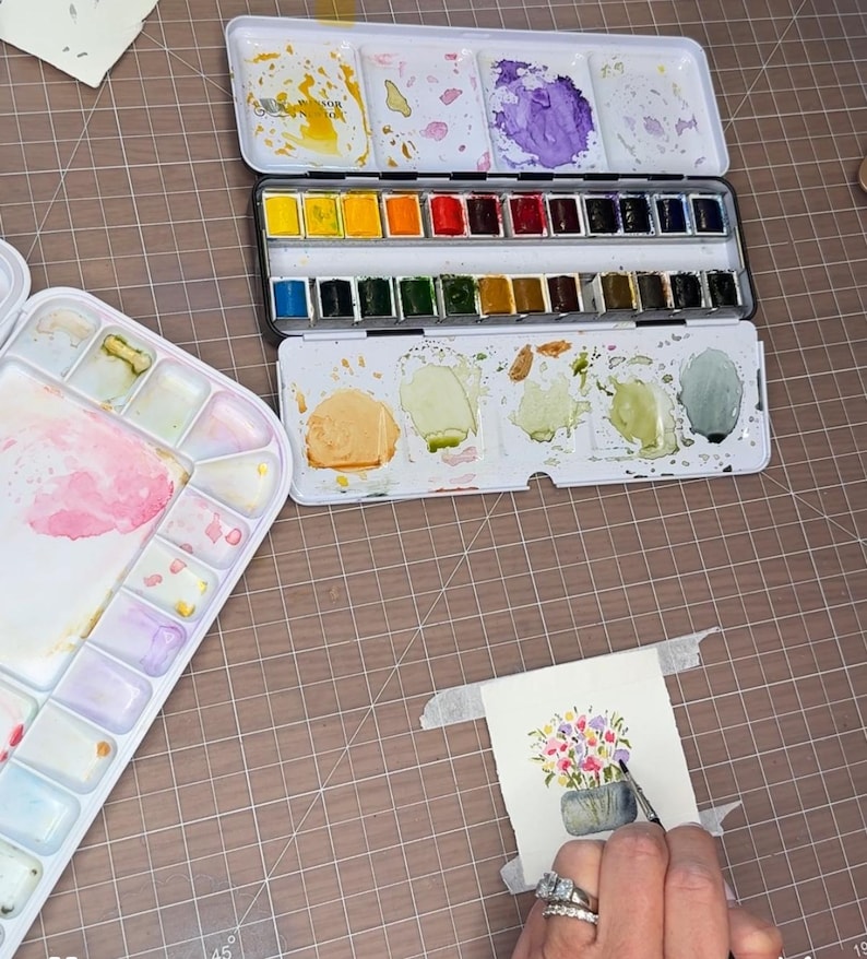 Pu&ograve; includere: Un'attrezzatura per la pittura ad acquerello con una tavolozza di metallo con vari colori ad acquerello e una tavolozza di plastica separata. Si sta lavorando a un piccolo dipinto ad acquerello di fiori in un vaso con un pennello. L'attrezzatura &egrave; su una superficie a griglia marrone.