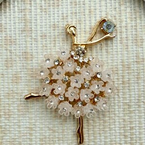 May include: A gold-toned ballerina brooch featuring a tutu made of translucent pale pink flower petals and clear rhinestones. The figure has a slender gold body, one arm raised with a single blue-tinted stone, and one leg extended.