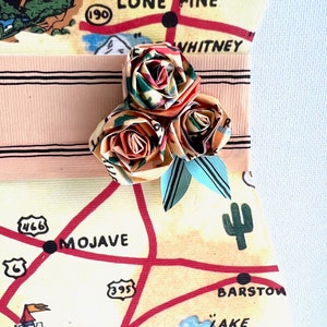 Pu&ograve; includere: Una composizione floreale di carta fatta a mano con tre rose origami nei toni dell'arancione, verde e rosso, accentuata da foglie di carta blu. I fiori sono attaccati a un nastro rosa chiaro su uno sfondo di mappa vintage con "LONE PINE" e "MOJAVE".