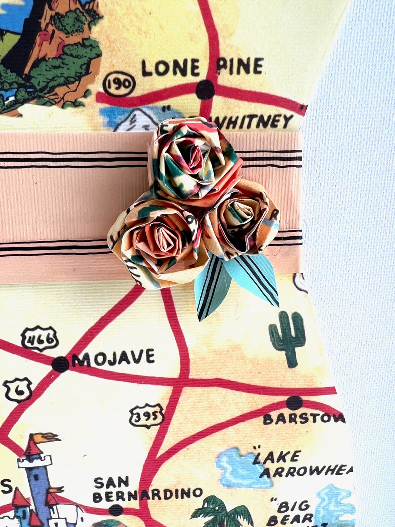 Pu&ograve; includere: Una composizione floreale di carta fatta a mano con tre rose origami nei toni dell'arancione, verde e rosso, accentuata da foglie di carta blu. I fiori sono attaccati a un nastro rosa chiaro su uno sfondo di mappa vintage con "LONE PINE" e "MOJAVE".