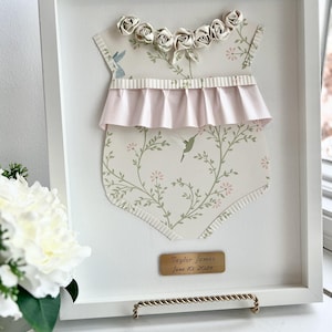 May include: A white frame with a white mat and a paper cutout of a baby onesie with a pink ruffle and floral pattern. The onesie has a row of paper roses around the neckline. The frame has a gold plaque with the text "Taylor James June 10, 2022".