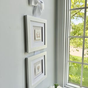 Pu&ograve; includere: Due quadri con cornice bianca con opere d'arte floreali sono montati su una parete azzurra. Un grande fiocco bianco &egrave; attaccato sopra la cornice superiore. A destra c'&egrave; una finestra con vista sul verde.