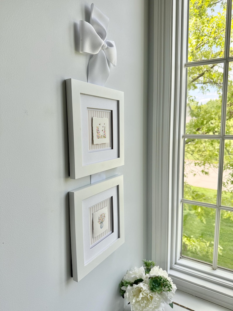 Pu&ograve; includere: Due quadri con cornice bianca con opere d'arte floreali sono montati su una parete azzurra. Un grande fiocco bianco &egrave; attaccato sopra la cornice superiore. A destra c'&egrave; una finestra con vista sul verde.