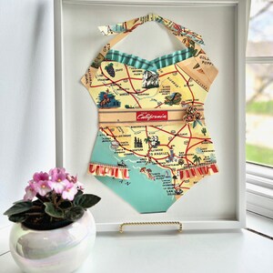 Pu&ograve; includere: Un'opera d'arte incorniciata a forma di costume da bagno, realizzata con una mappa vintage della California. Sono visibili il collo all'americana e i bordi a balze. L'opera d'arte &egrave; esposta su una superficie bianca, con una piccola pianta in vaso e una finestra sullo sfondo.
