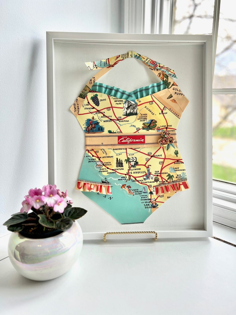 Pu&ograve; includere: Un'opera d'arte incorniciata a forma di costume da bagno, realizzata con una mappa vintage della California. Sono visibili il collo all'americana e i bordi a balze. L'opera d'arte &egrave; esposta su una superficie bianca, con una piccola pianta in vaso e una finestra sullo sfondo.