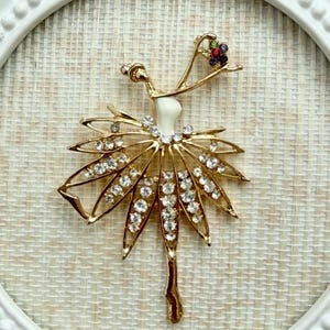 May include: A gold-toned ballerina brooch featuring a white enamel torso, a skirt adorned with clear rhinestones, and a small cluster of multicolored gemstones at the head. The figure is depicted in a dance pose with one arm raised.