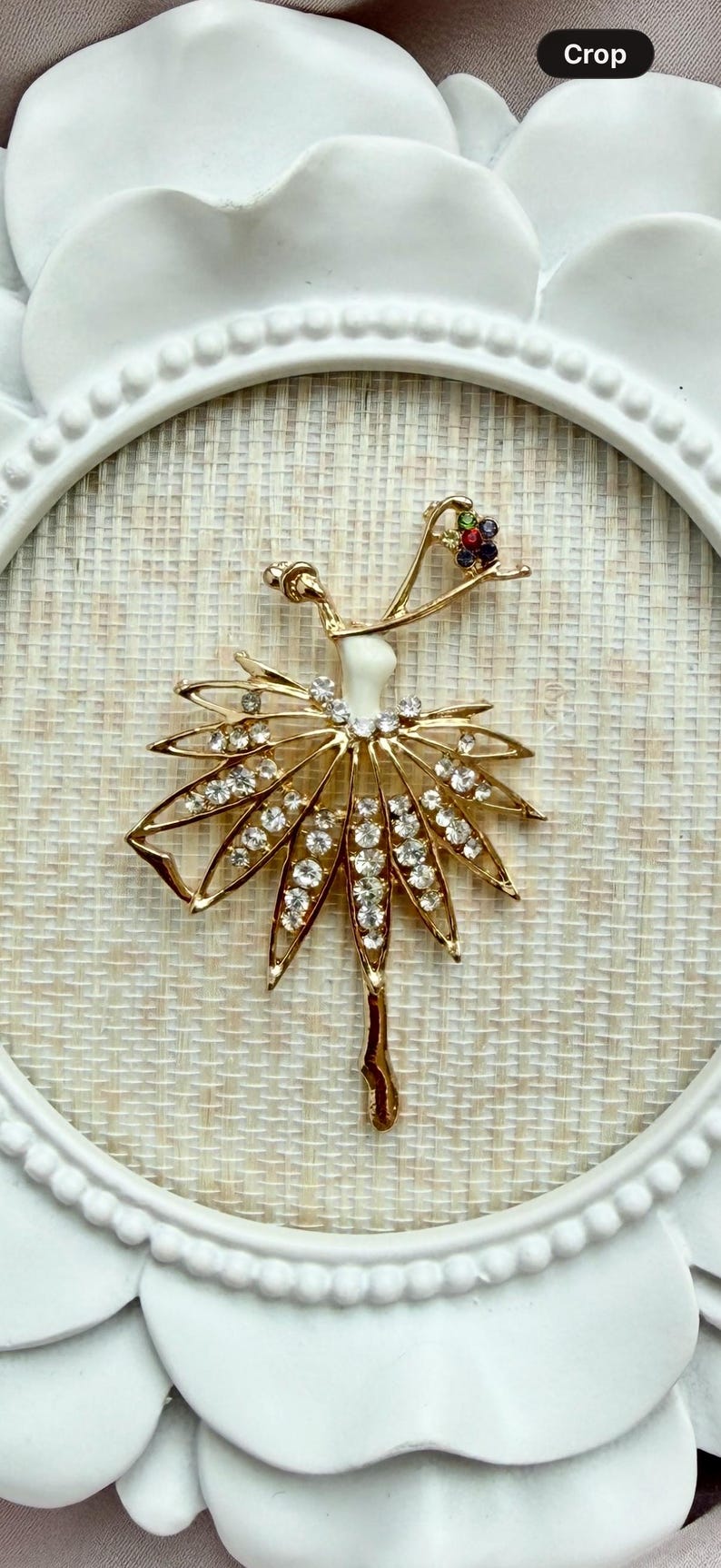 May include: A gold-toned ballerina brooch featuring a white enamel torso, a skirt adorned with clear rhinestones, and a small cluster of multicolored gemstones at the head. The figure is depicted in a dance pose with one arm raised.