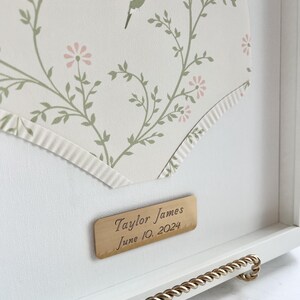 May include: A white frame with a gold trim and a gold plaque that reads "Taylor James June 10, 2024". The frame contains a white background with a floral pattern and a small hummingbird.